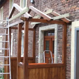 Drewniana konstrukcja ganku w trakcie budowy, widoczne elementy szkieletu i drabina oparta o ścianę budynku z czerwonej cegły. Częściowo pomalowane elementy konstrukcji.