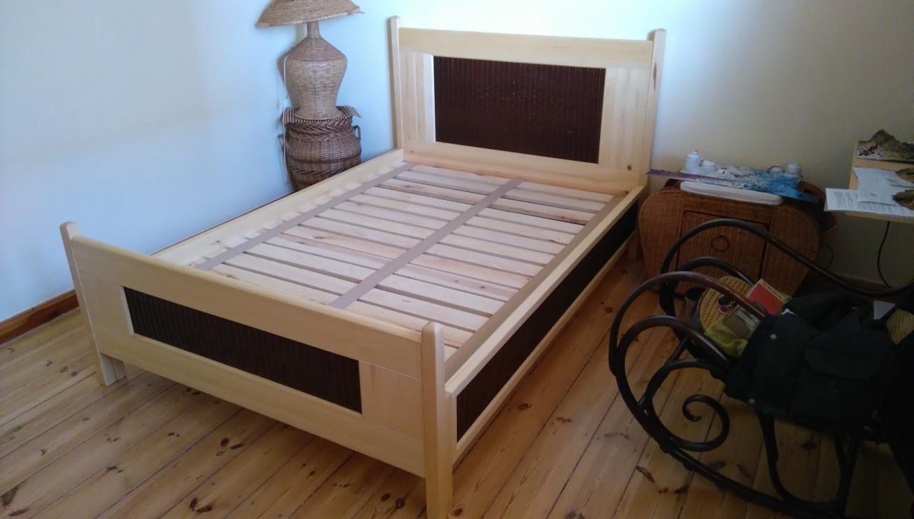 Drewniane łóżko z jasnego drewna z ciemnobrązowymi wstawkami na wezgłowiu i w nogach, widoczny stelaż pod materac, w tle lampa z wikliny i drewniana podłoga.