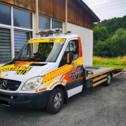 Biała laweta Mercedes Sprinter z pomarańczowymi akcentami i logotypem 'Assistance 24/7' zaparkowana na żwirowym podłożu przed budynkiem z dużymi oknami.