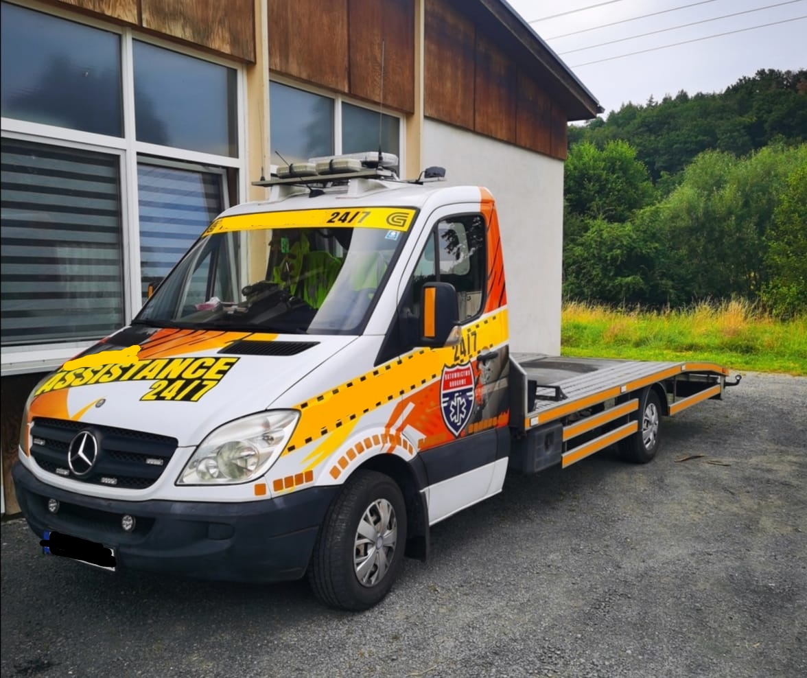 Biała laweta Mercedes Sprinter z pomarańczowymi akcentami i logotypem 'Assistance 24/7' zaparkowana na żwirowym podłożu przed budynkiem z dużymi oknami.