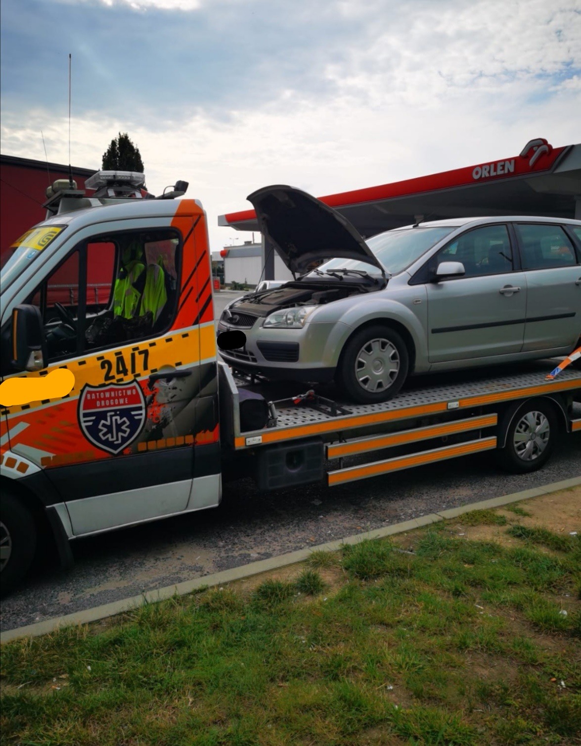 Srebrny Ford Focus z otwartą maską na lawecie z oznaczeniami 'Ratownictwo Drogowe 24/7' na tle stacji Orlen.