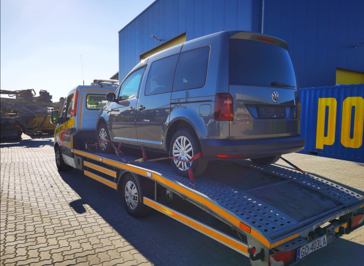 Szara laweta z Volkswagenem Caddy zabezpieczonym pasami transportowymi, na tle błękitnego magazynu i sprzętu budowlanego.
