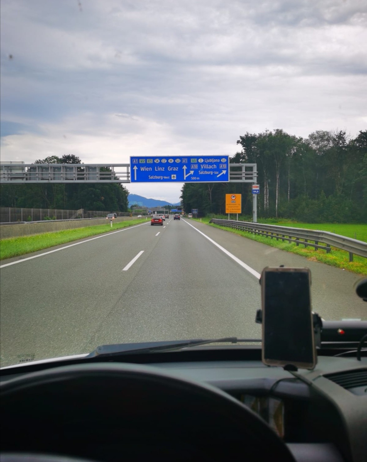 Widok z kabiny pojazdu na autostradę A1 w Austrii, kierunek na Wiedeń, Linz i Graz, z tablicą informacyjną nad drogą i uchwytem na telefon widocznym na desce rozdzielczej.