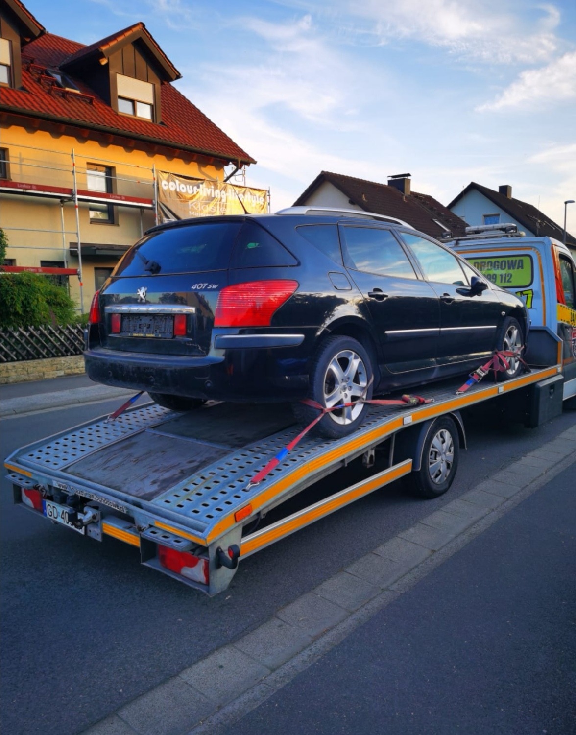 Czarny samochód Peugeot 407 SW zabezpieczony czerwonymi pasami na platformie lawety na tle zabudowań mieszkalnych i błękitnego nieba.