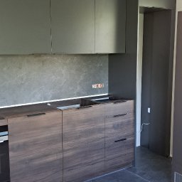 Firma WKTM - Szare szafki kuchenne podwieszane z ciemnobrązowymi szafkami dolnymi o drewnianej fakturze, oświetlenie szynowe, szara ściana nad blatem, widoczne gniazdka elektryczne, podłoga w trakcie wykańczania.