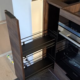 Firma WKTM - Wysuwana szafka cargo z dwoma poziomami półek w zabudowie kuchennej z forniru w odcieniach brązu, obok piekarnik Siemens, widoczne niedoskonałości wykończenia podłogi.