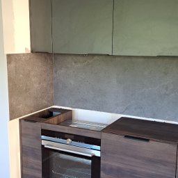 Firma WKTM - Montaż kuchni: szafki wiszące w kolorze grafitowym, zabudowa piekarnika Siemens w fornirze drewnopodobnym, szara ściana między szafkami, widoczne niedokończone prace wykończeniowe.
