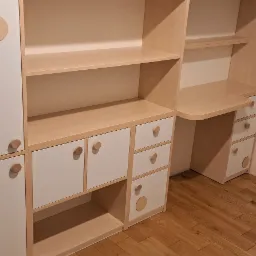 Biało-beżowy regał z biurkiem w pokoju dziecięcym, okrągłe uchwyty i dekoracyjne kółka, drewniana podłoga.