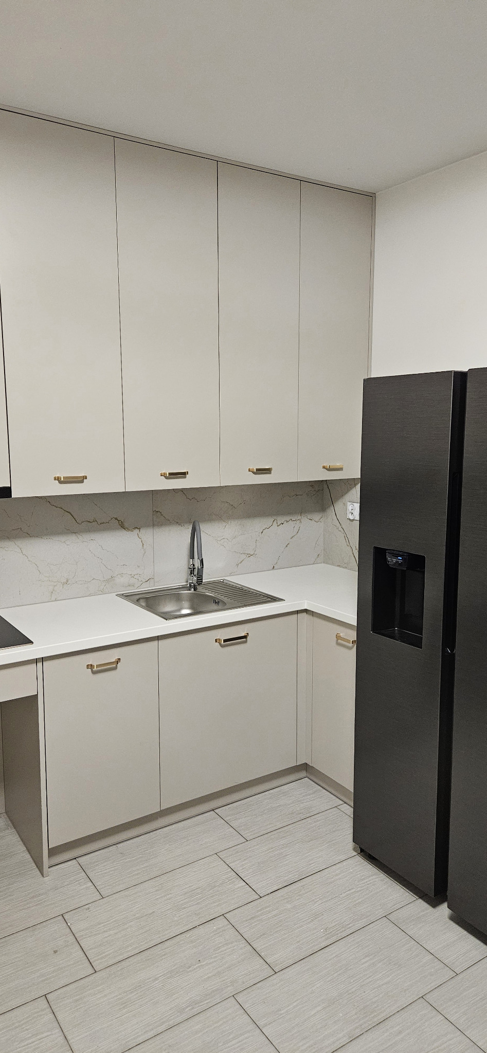 Jasna kuchnia z górnymi i dolnymi szafkami z beżowymi frontami, złotymi uchwytami, marmurowym backsplashiem i czarną lodówką z dozownikiem wody.