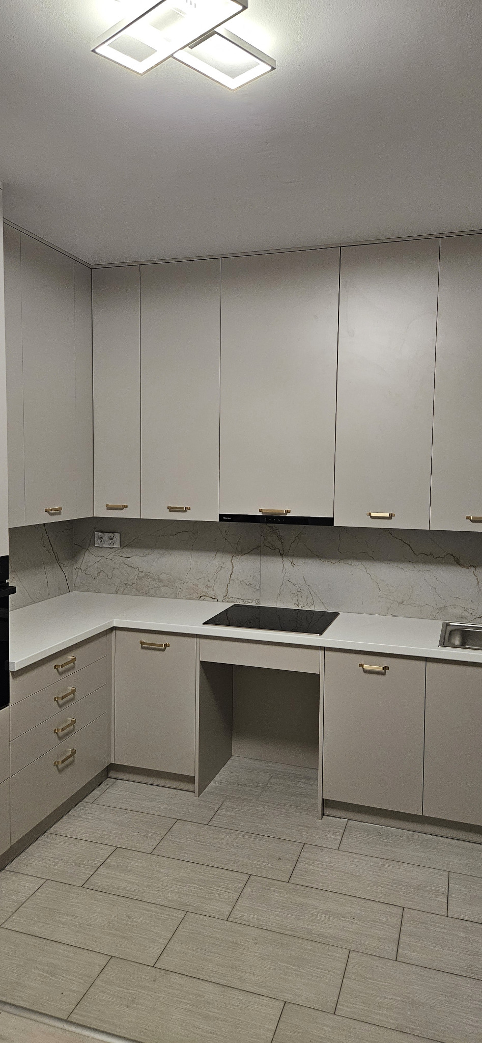 Jasnoszara kuchnia z frontami bez uchwytów, złotymi relingami i blatem imitującym marmur, oświetlona nowoczesną lampą sufitową.