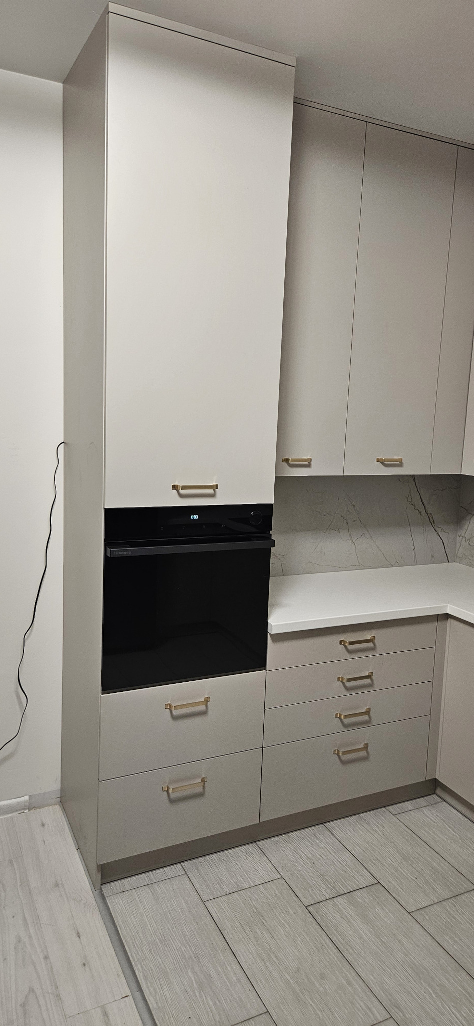 Szara zabudowa kuchenna z piekarnikiem i złotymi uchwytami, marmurowy panel nad blatem, minimalistyczny design.