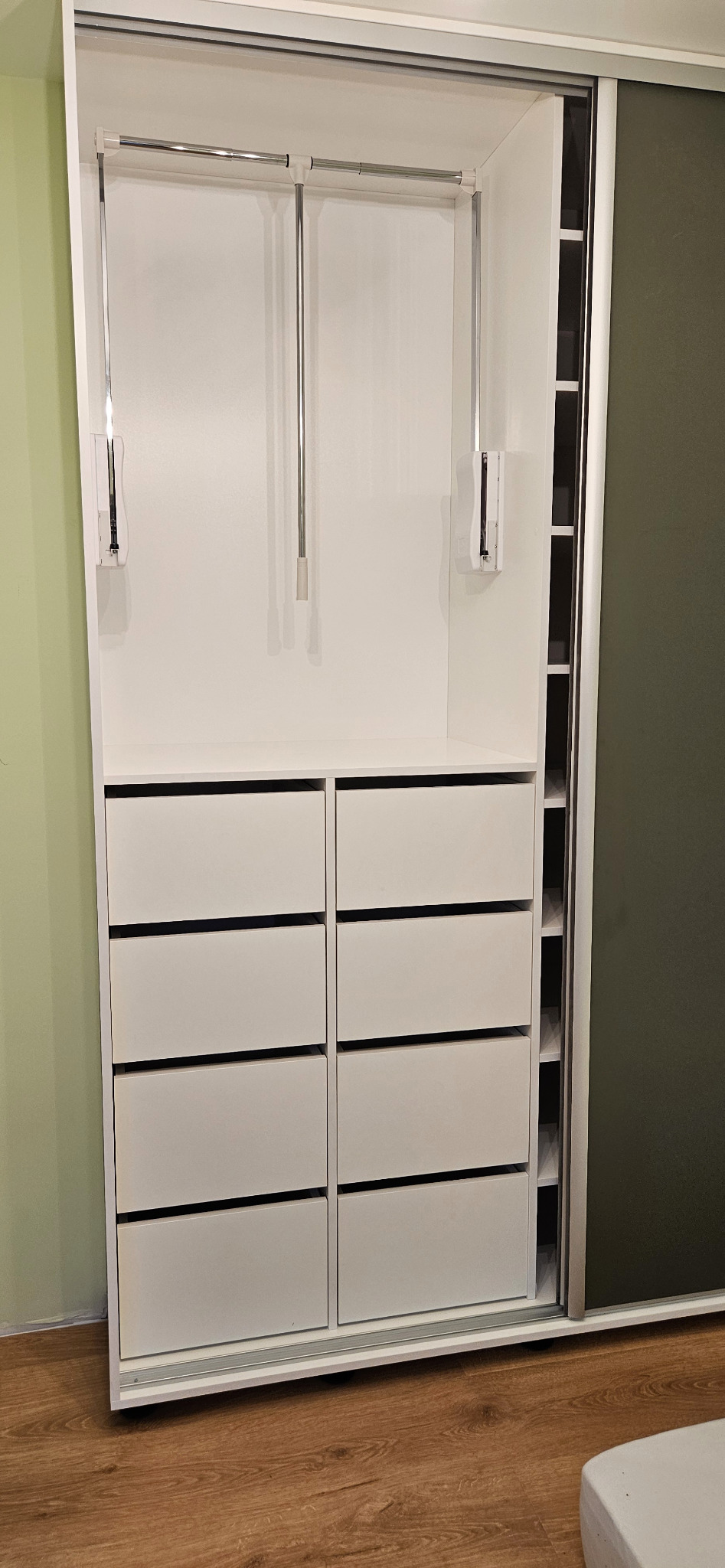 Biała szafa przesuwna z rzędami szuflad i metalowymi drążkami na ubrania, widoczna częściowo zasunięta ciemna płyta drzwi przesuwnych.