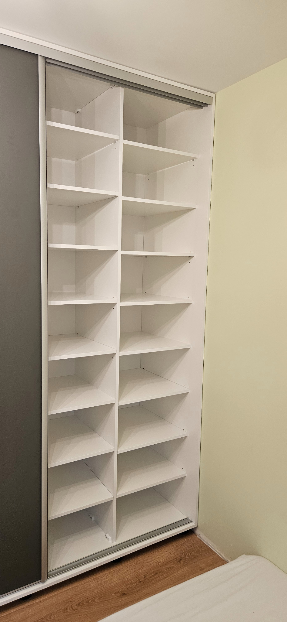 Biała szafa wnękowa z przesuwnymi drzwiami, widok na puste półki, jedna z szarych płyt drzwiowych zasunięta.