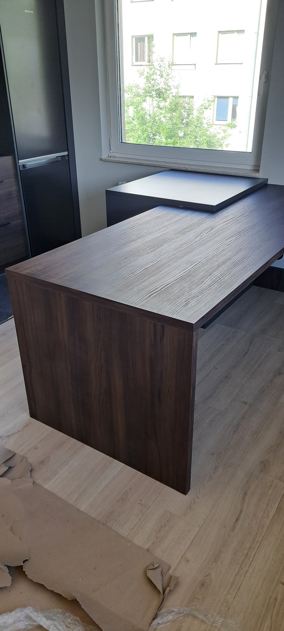 Kątowe biurko z blatem w kolorze jasnego drewna i ciemnobrązową podstawą, widok z boku na tle okna i szafy, z resztkami opakowania na podłodze.