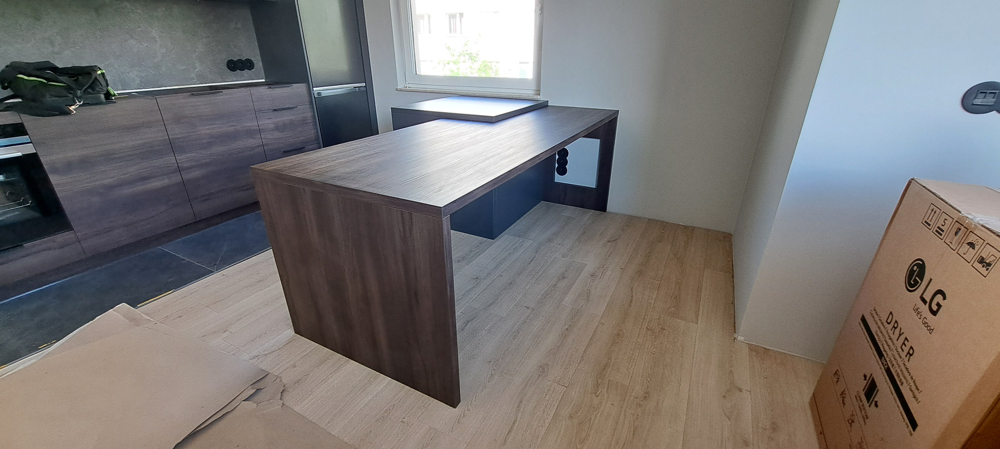 Nowoczesna kuchnia z drewnianymi szafkami i blatem roboczym zintegrowanym z oknem, widoczny karton z nowym sprzętem AGD.