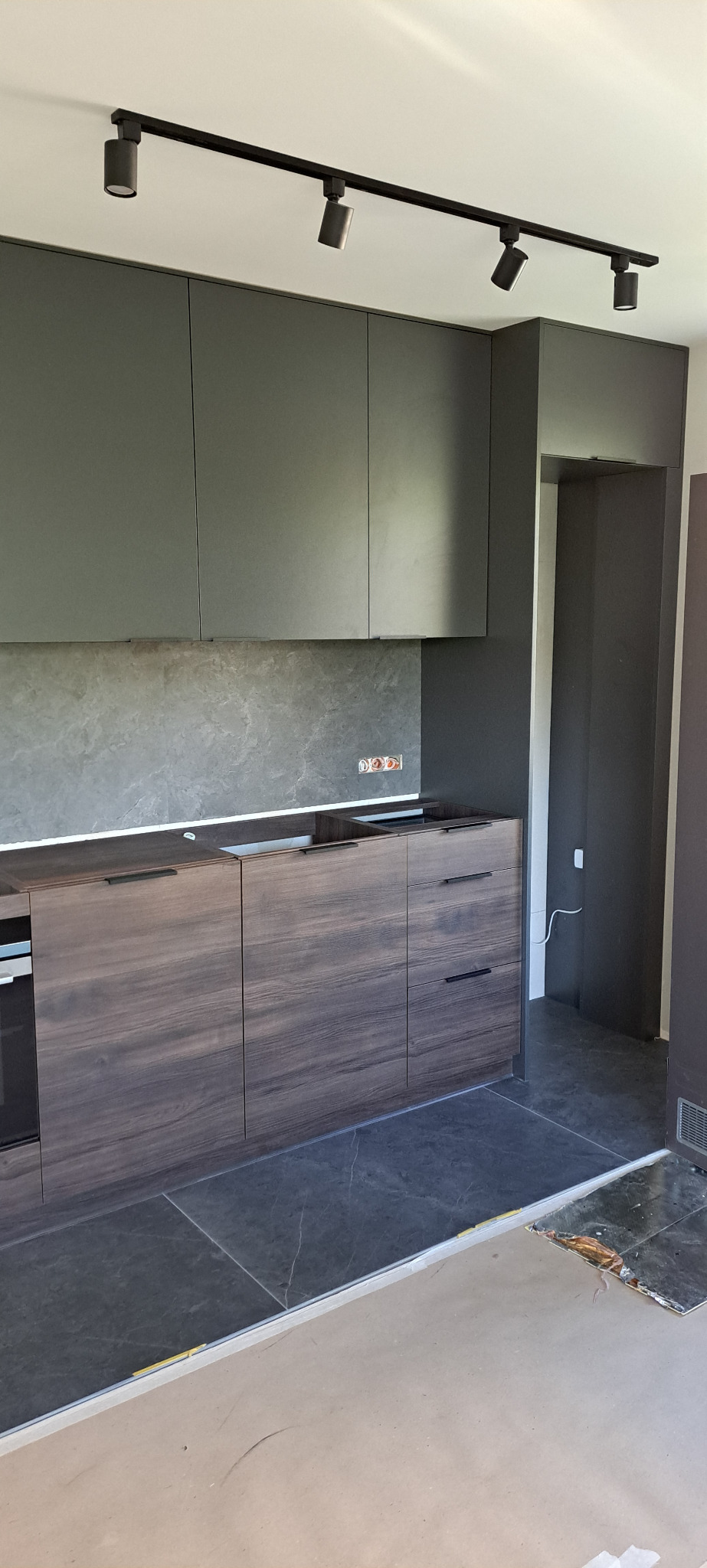 Szare szafki kuchenne podwieszane z ciemnobrązowymi szafkami dolnymi o drewnianej fakturze, oświetlenie szynowe, szara ściana nad blatem, widoczne gniazdka elektryczne, podłoga w trakcie wykańczania.