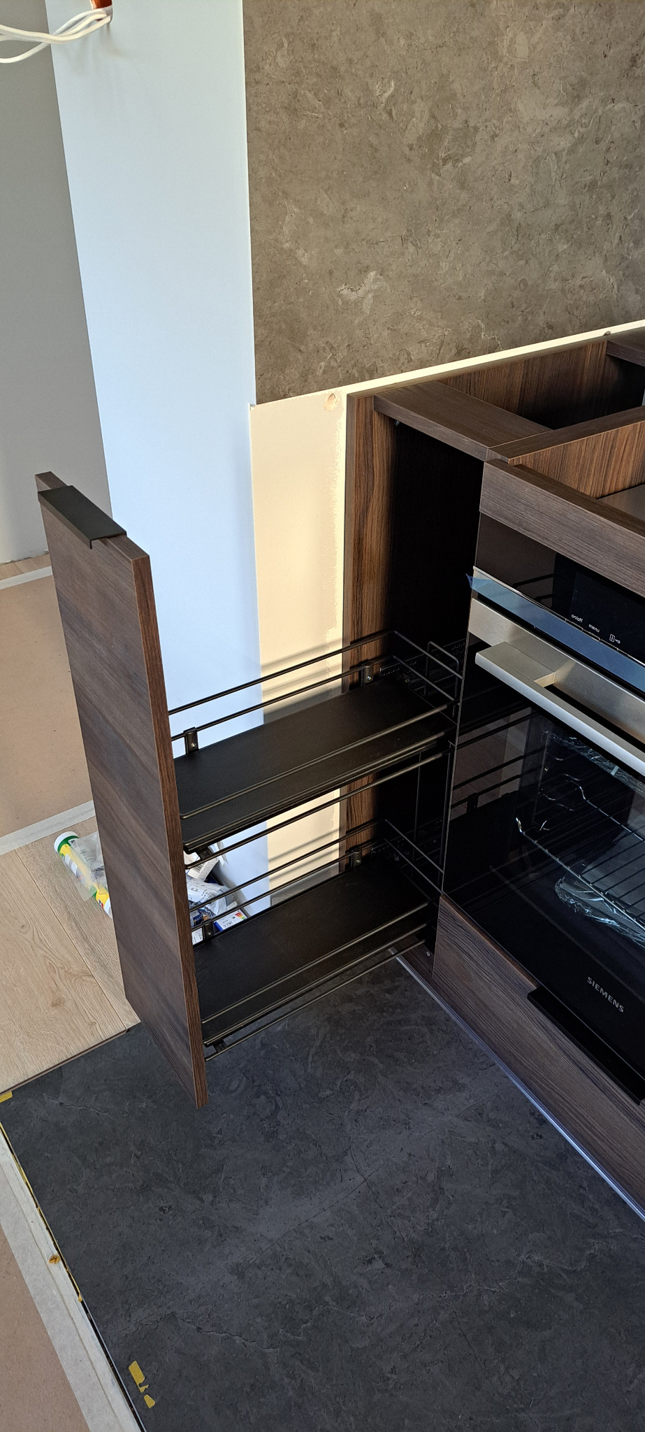 Wysuwana szafka cargo z dwoma poziomami półek w zabudowie kuchennej z forniru w odcieniach brązu, obok piekarnik Siemens, widoczne niedoskonałości wykończenia podłogi.