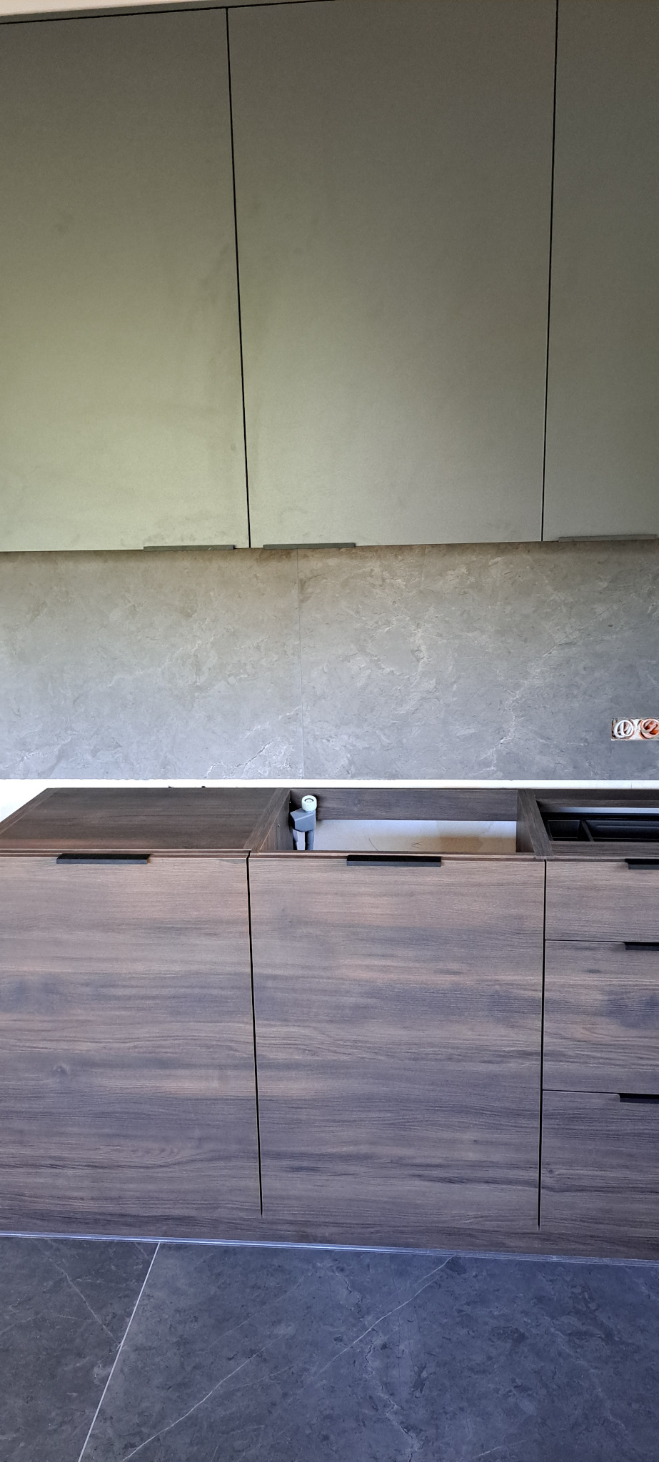 Nowoczesna zabudowa kuchenna z ciemnego drewna i szarych szafek wiszących, widoczny fragment instalacji hydraulicznej w miejscu zlewu.