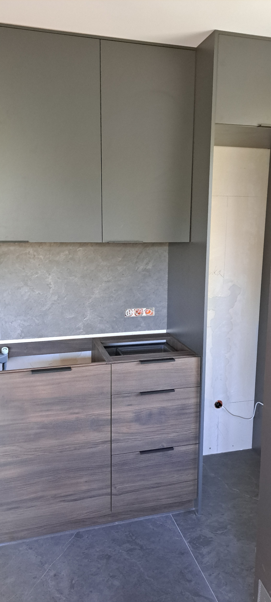 Szare szafki kuchenne z drewnopodobnym frontem, widoczne gniazdka elektryczne w ścianie i nieukończona instalacja elektryczna w tle.