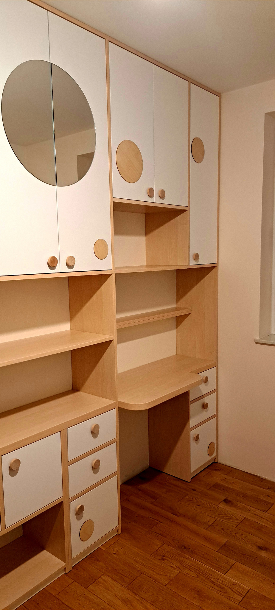 Biało-drewniana zabudowa meblowa z okrągłymi uchwytami i okrągłym lustrem, obejmująca szafy, półki i biurko, na drewnianej podłodze.