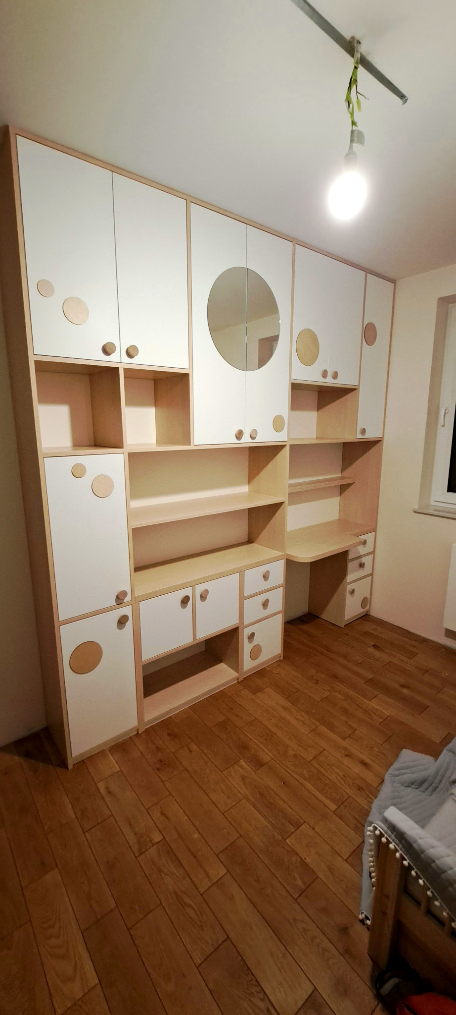 Zabudowa meblowa z biurkiem, szafkami i półkami w pokoju dziecięcym, fronty białe z okrągłymi dekorami, drewniane blaty i uchwyty, okrągłe lustro na jednej z szafek górnych.