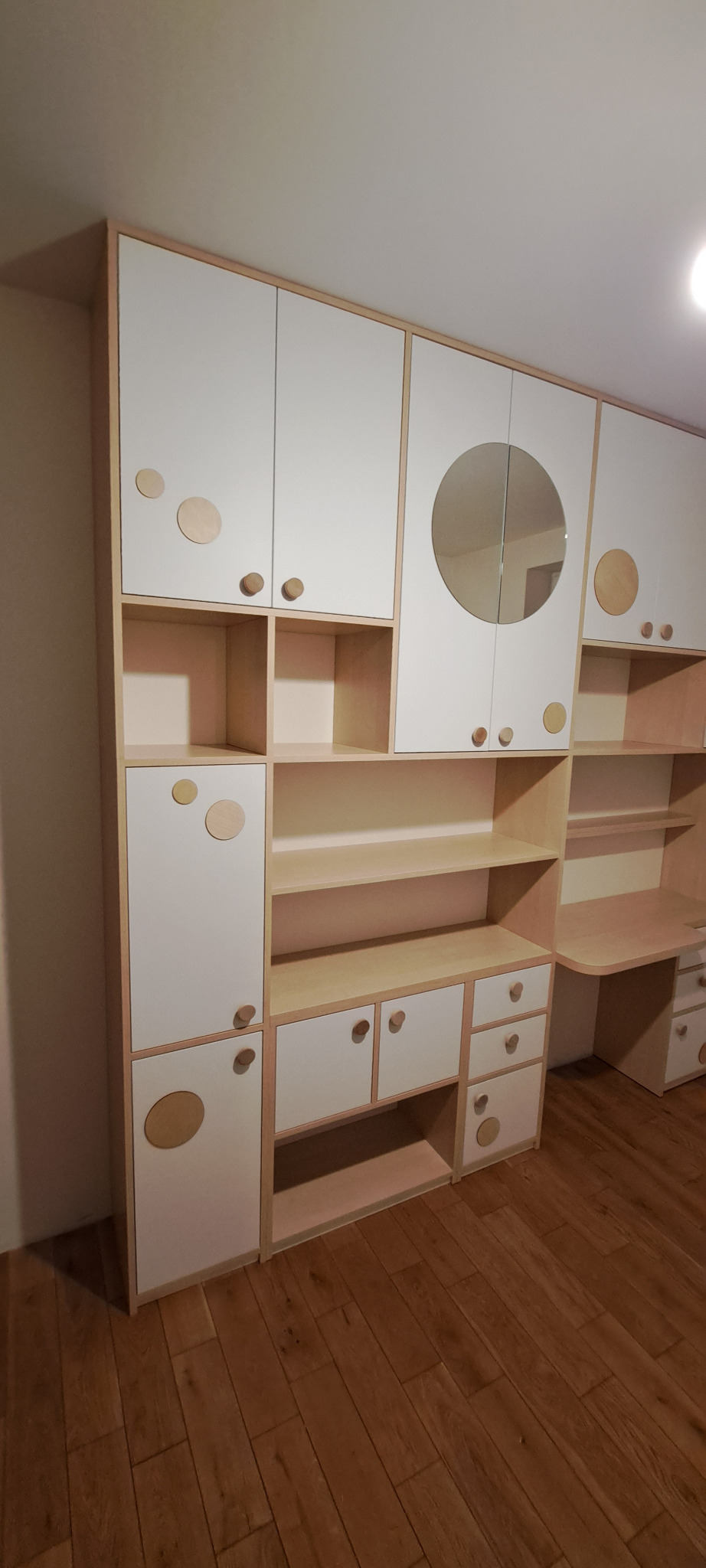 Biało-beżowa zabudowa meblowa z okrągłymi dekoracjami i okrągłym lustrem, w pokoju dziecięcym, z biurkiem po prawej stronie.