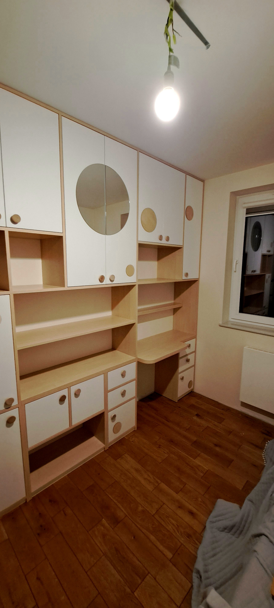 Zabudowa meblowa dziecięca z biurkiem, fronty białe z drewnianymi okrągłymi uchwytami, widoczne również okrągłe dekoracje na frontach, podłoga drewniana.