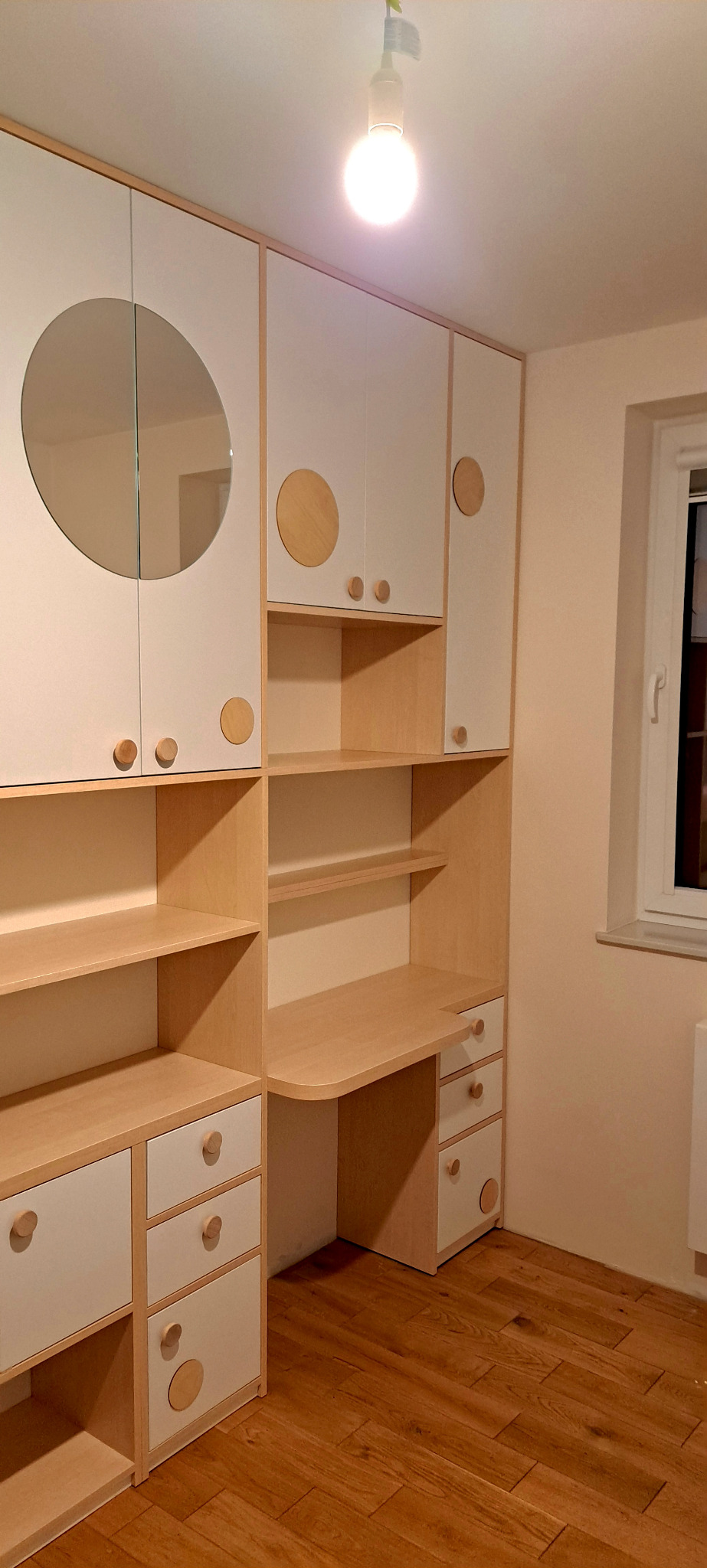 Biało-drewniana zabudowa meblowa z okrągłym lustrem i biurkiem, widoczna w pokoju z drewnianą podłogą, oświetlona żarówką.