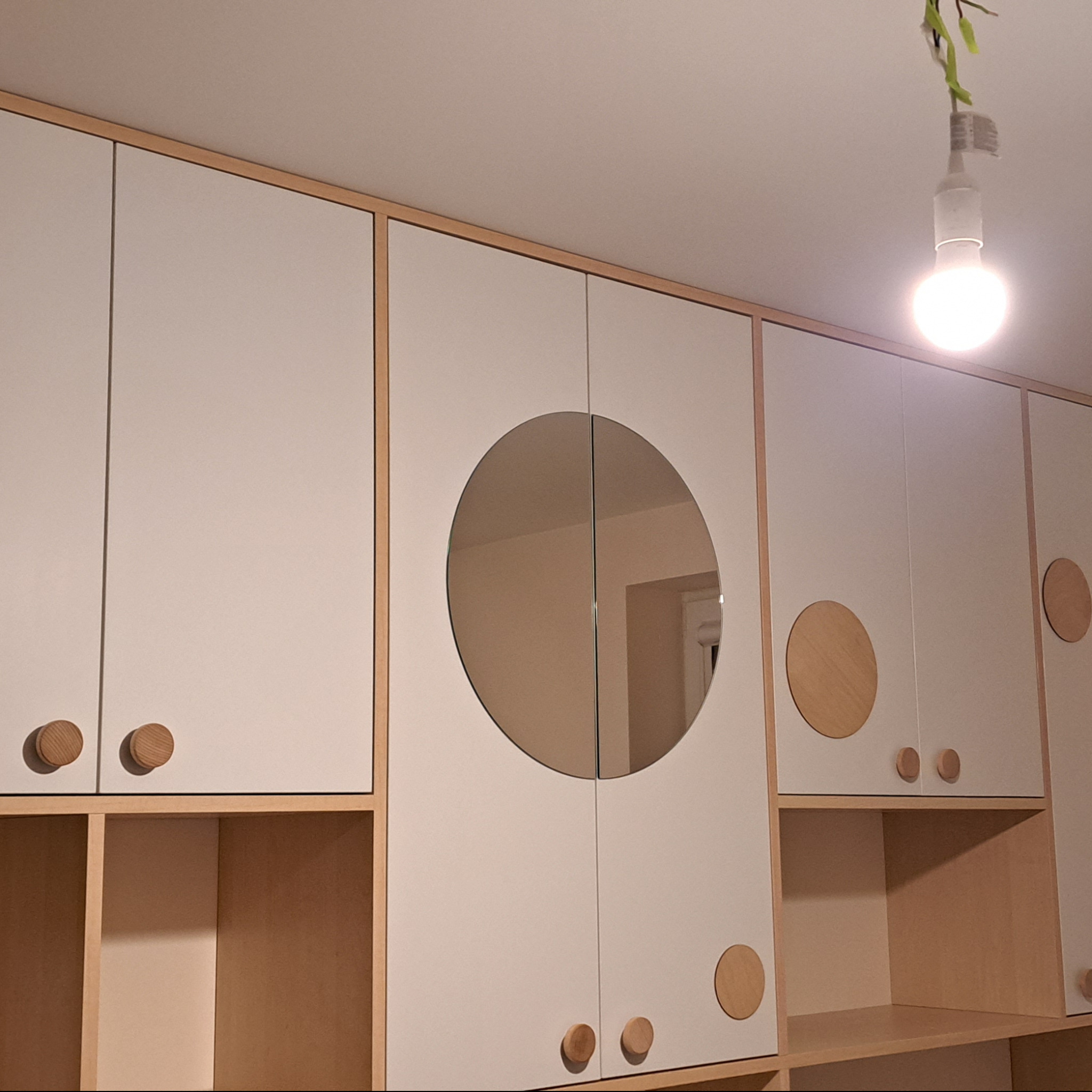 Biała szafa na wymiar z okrągłym lustrem i drewnianymi uchwytami, wykonana w Krakowie, z widocznym oświetleniem sufitowym.
