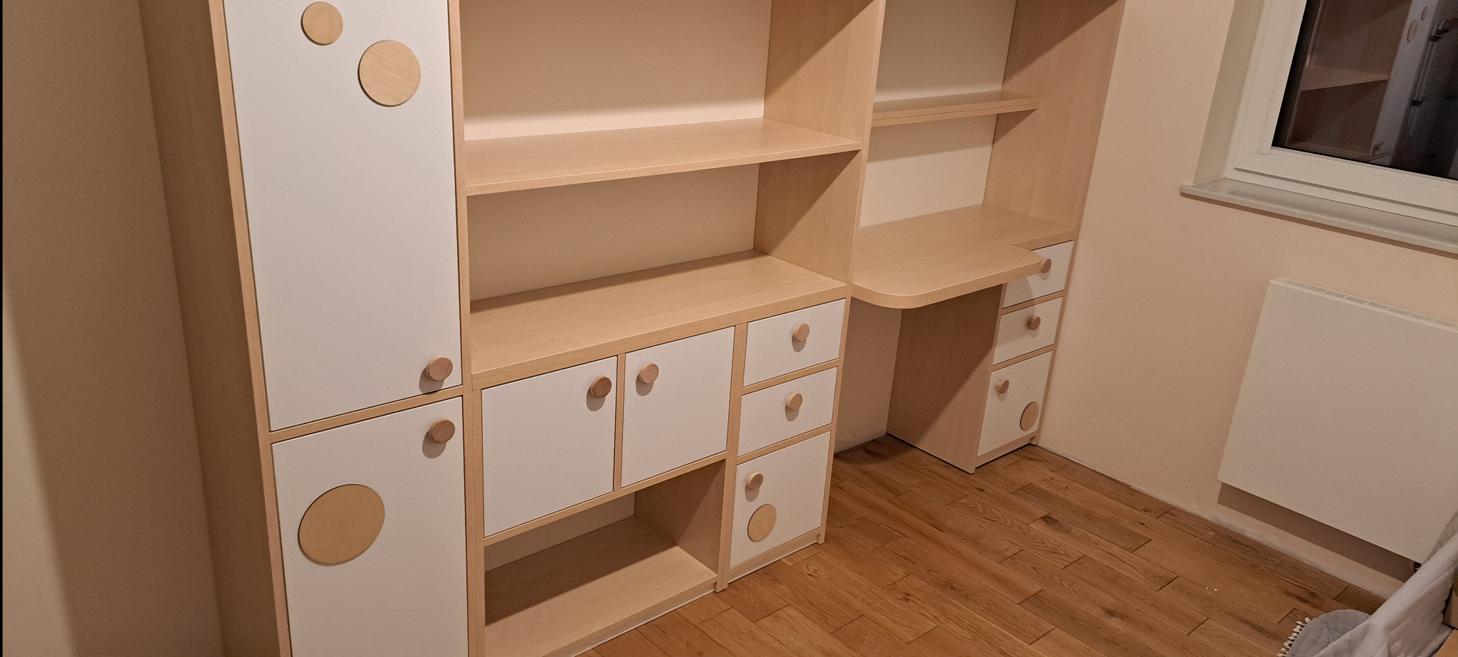 Biało-beżowy regał z biurkiem w pokoju dziecięcym, okrągłe uchwyty i dekoracyjne kółka, drewniana podłoga.