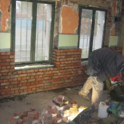 Mężczyzna w trakcie murowania ściany z cegły przy oknie w surowym wnętrzu. Widoczne narzędzia i materiały budowlane, ściana częściowo otynkowana.