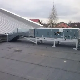 Szara centrala wentylacyjna VTS na dachu budynku z szarym niebem w tle. Widoczna metalowa konstrukcja wsporcza i kanały wentylacyjne. Ujęcie z boku.