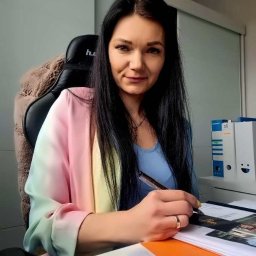 BOSS SYLWIA BOKSA - Kobieta z długimi czarnymi włosami siedzi przy biurku, trzyma długopis nad dokumentami i patrzy w obiektyw. Na biurku leżą projekty budynków i segregatory.