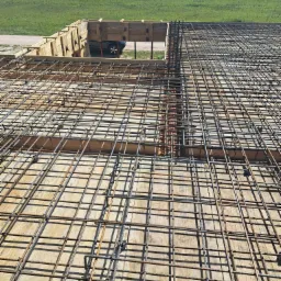 Zbrojenie płyty fundamentowej z deskowaniem, widok z góry na siatkę stalową i drewniane szalunki, w tle zielony teren z drzewami.