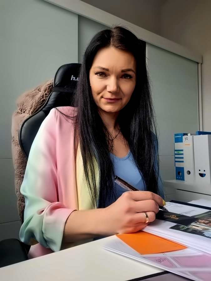 Kobieta z długimi czarnymi włosami siedzi przy biurku, trzyma długopis nad dokumentami i patrzy w obiektyw. Na biurku leżą projekty budynków i segregatory.