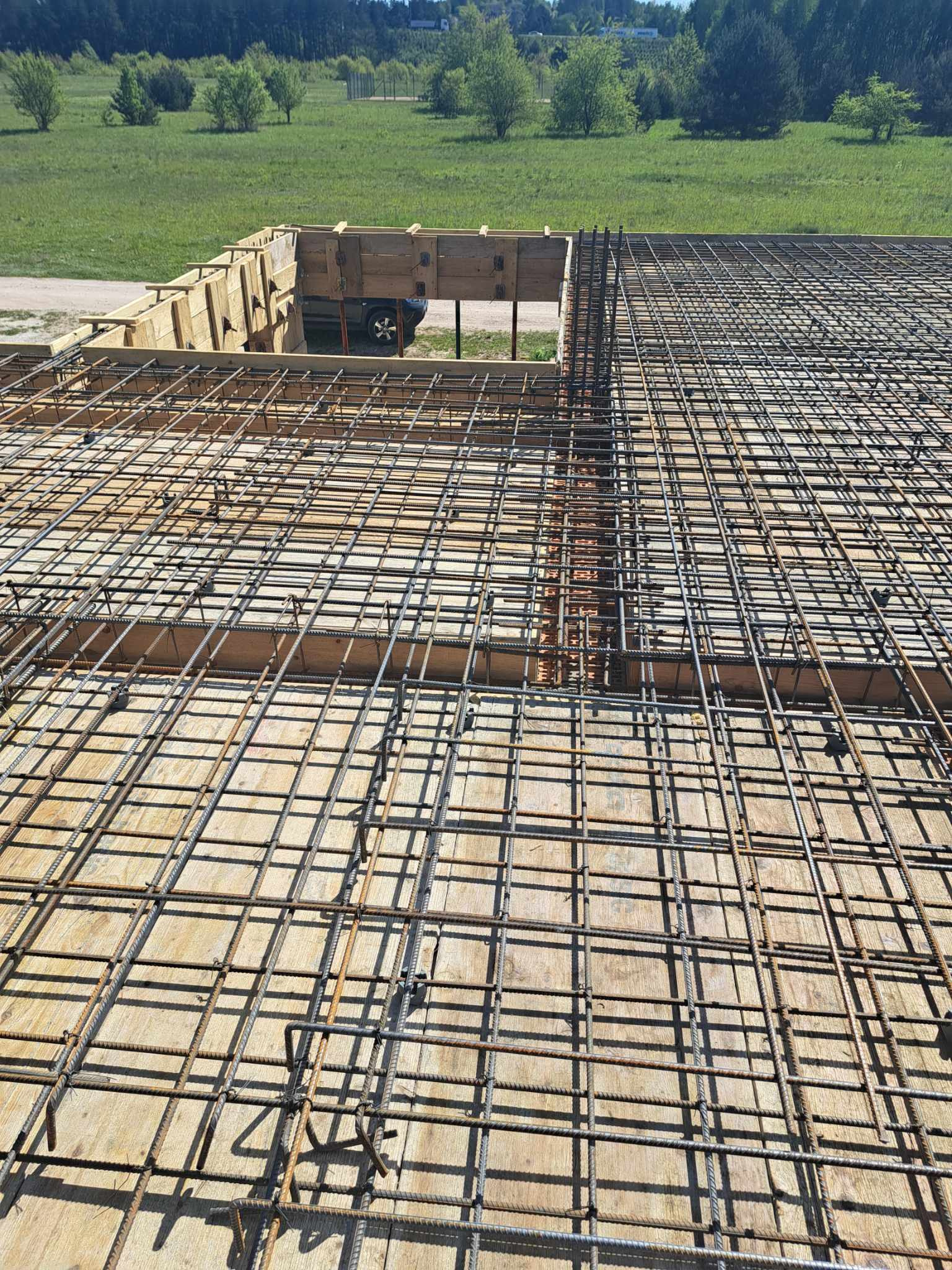 Zbrojenie płyty fundamentowej z deskowaniem, widok z góry na siatkę stalową i drewniane szalunki, w tle zielony teren z drzewami.