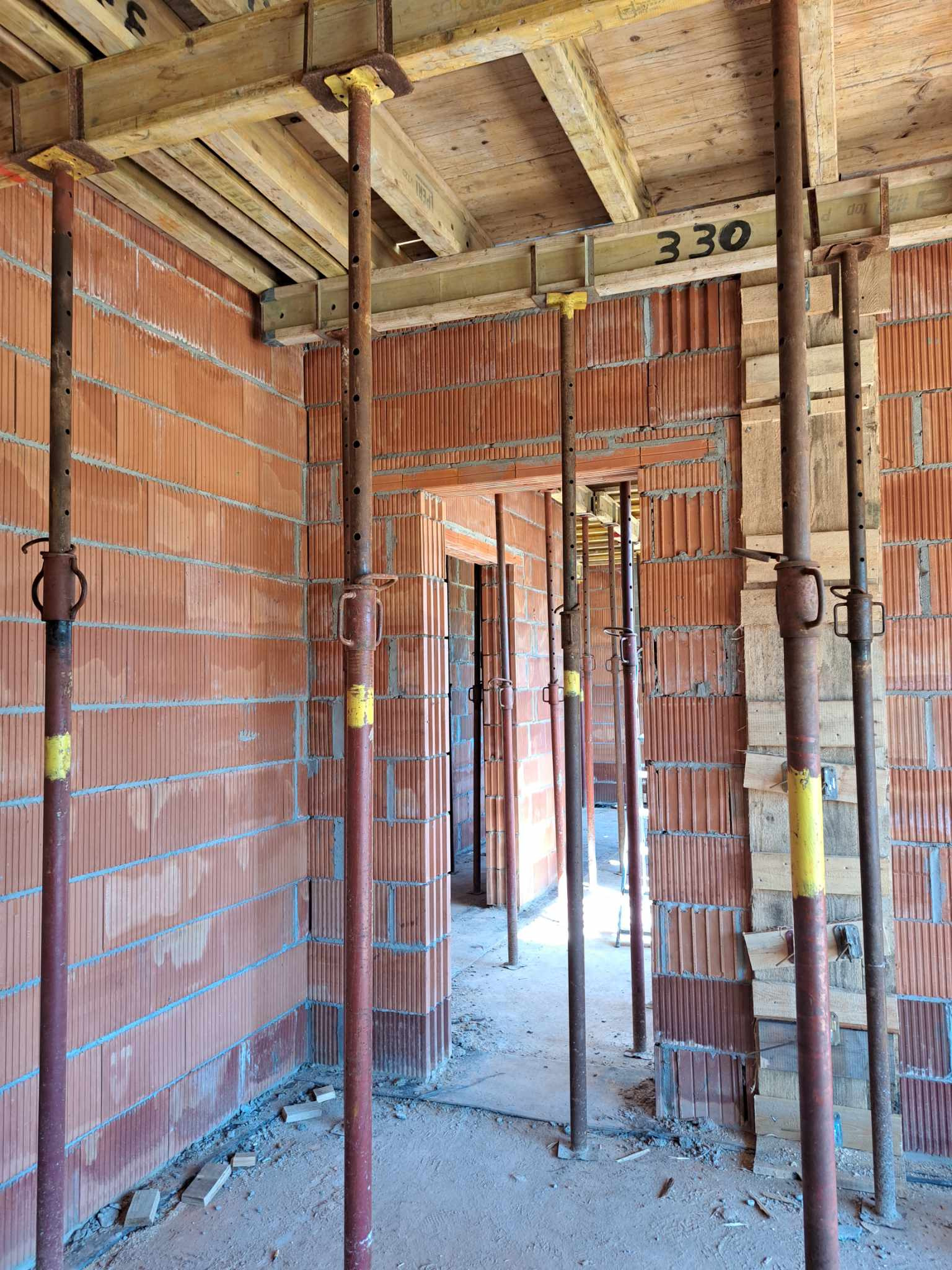 Wnętrze surowego budynku w trakcie budowy, z widocznymi ścianami z czerwonej cegły dziurawki, metalowymi stemplami budowlanymi z żółtymi elementami podtrzymującymi drewniane szalunki stropu...