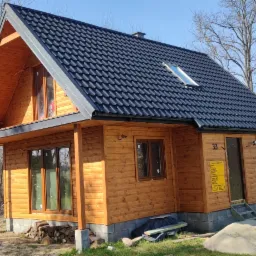 Drewniany dom letniskowy z czarną, skośną dachówką, widok z zewnątrz, w tle drzewa bez liści i błękitne niebo.