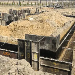 Przygotowanie wykopu pod fundamenty z szalunkami metalowymi i zbrojeniem stalowym, widok na plac budowy z ziemią i drzewami w tle.