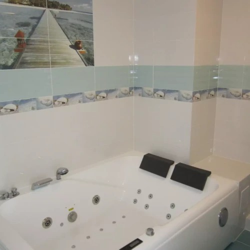 Biała wanna z hydromasażem w łazience z jasnymi płytkami i dekoracyjnym pasem z motywami morskimi oraz obrazem plaży z drewnianym pomostem.