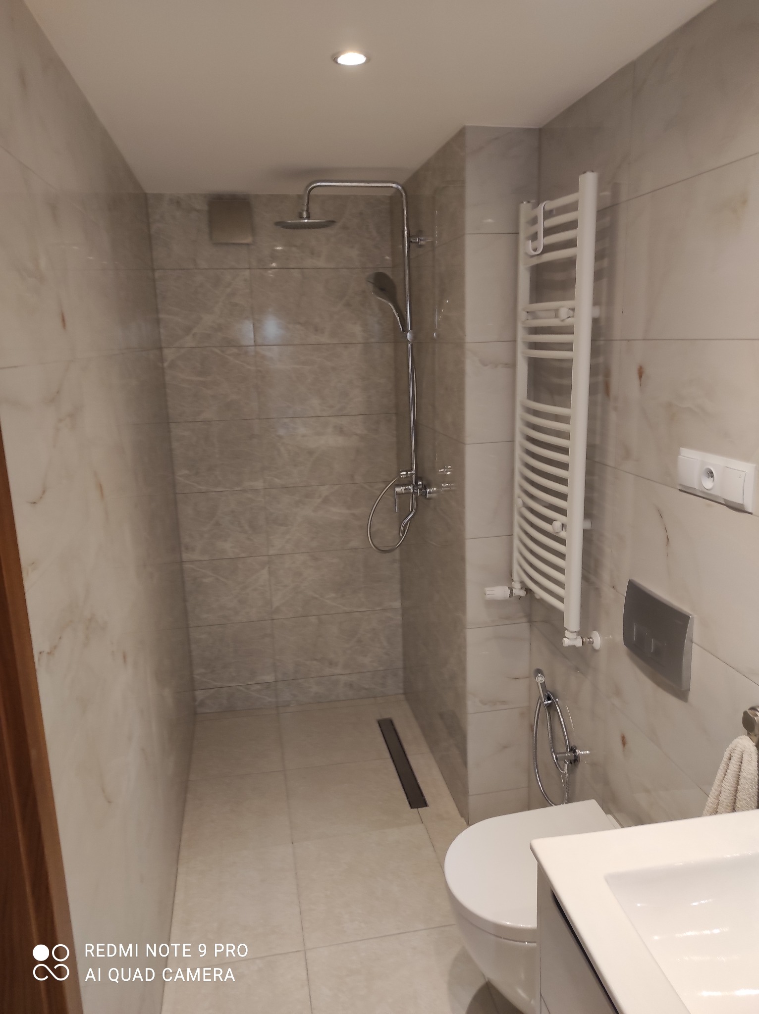 Nowoczesna łazienka z marmurowymi płytkami, kabiną prysznicową typu walk-in, grzejnikiem drabinkowym i podtynkową spłuczką.