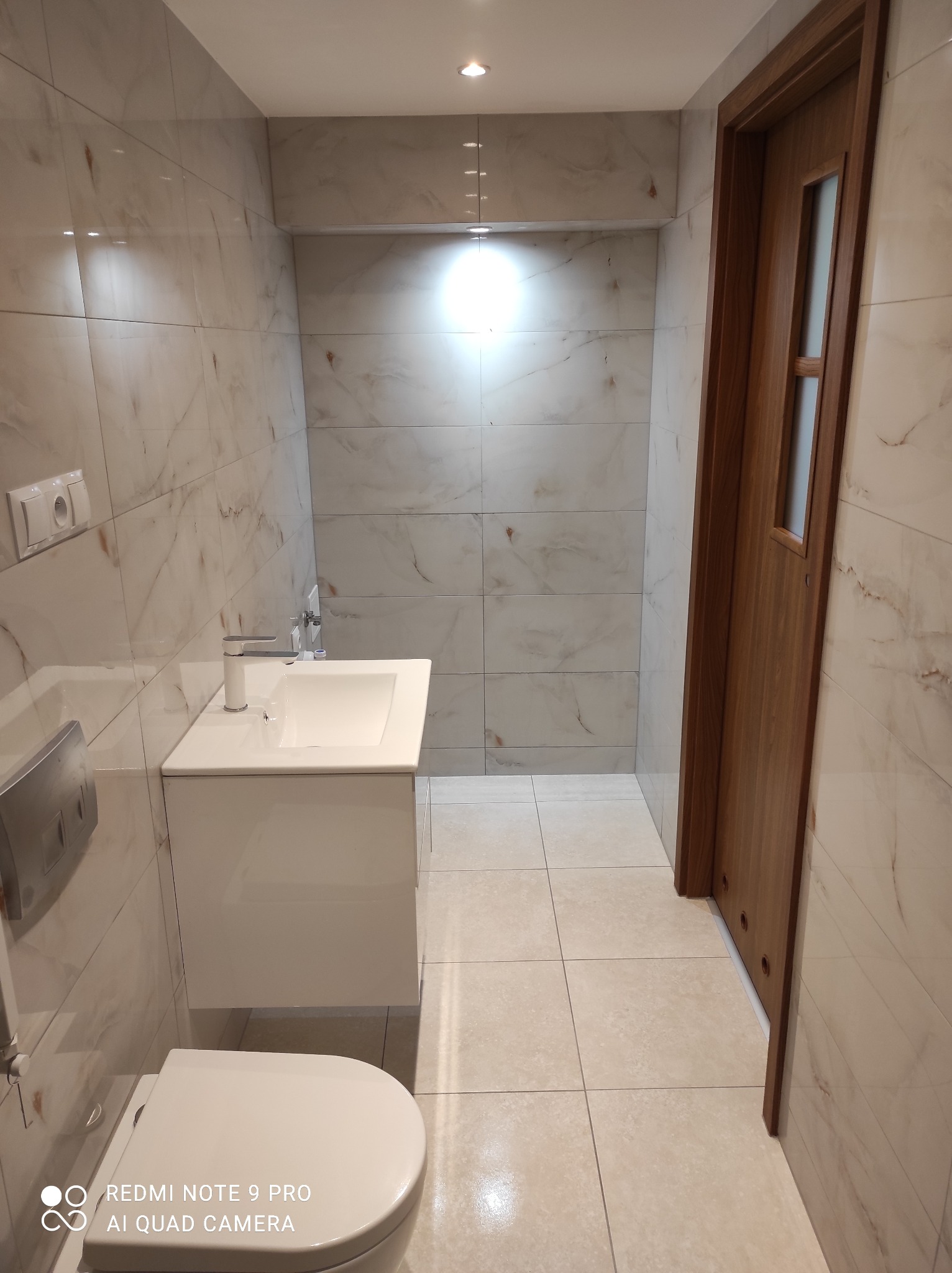 Nowoczesna łazienka z białymi marmurkowymi płytkami, wiszącą toaletą, umywalką z szafką i drewnianymi drzwiami.