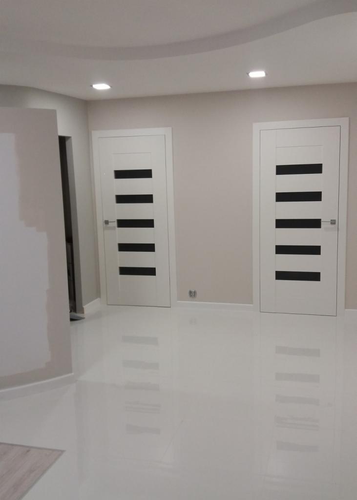 Dwa białe, nowoczesne drzwi wewnętrzne z czarnymi, poziomymi paskami, zamontowane w jasnym pomieszczeniu z połyskującą, białą podłogą.