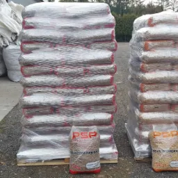 Dwa palety pelletu drzewnego owinięte folią, z widocznymi workami 15kg z logo PSP na pierwszym planie. W tle widać duże worki i fragment ciężarówki.