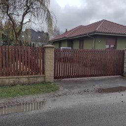 Erc Technik - Drewniana brama wjazdowa z pionowych desek, osadzona na murowanych słupkach, przed domem z czerwoną dachówką, widok na kałużę na asfalcie.