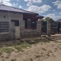 Nowoczesne ogrodzenie panelowe z elementami imitującymi kamień, skrzynka elektryczna na słupku, dom jednorodzinny w tle, widok z poziomu gruntu.