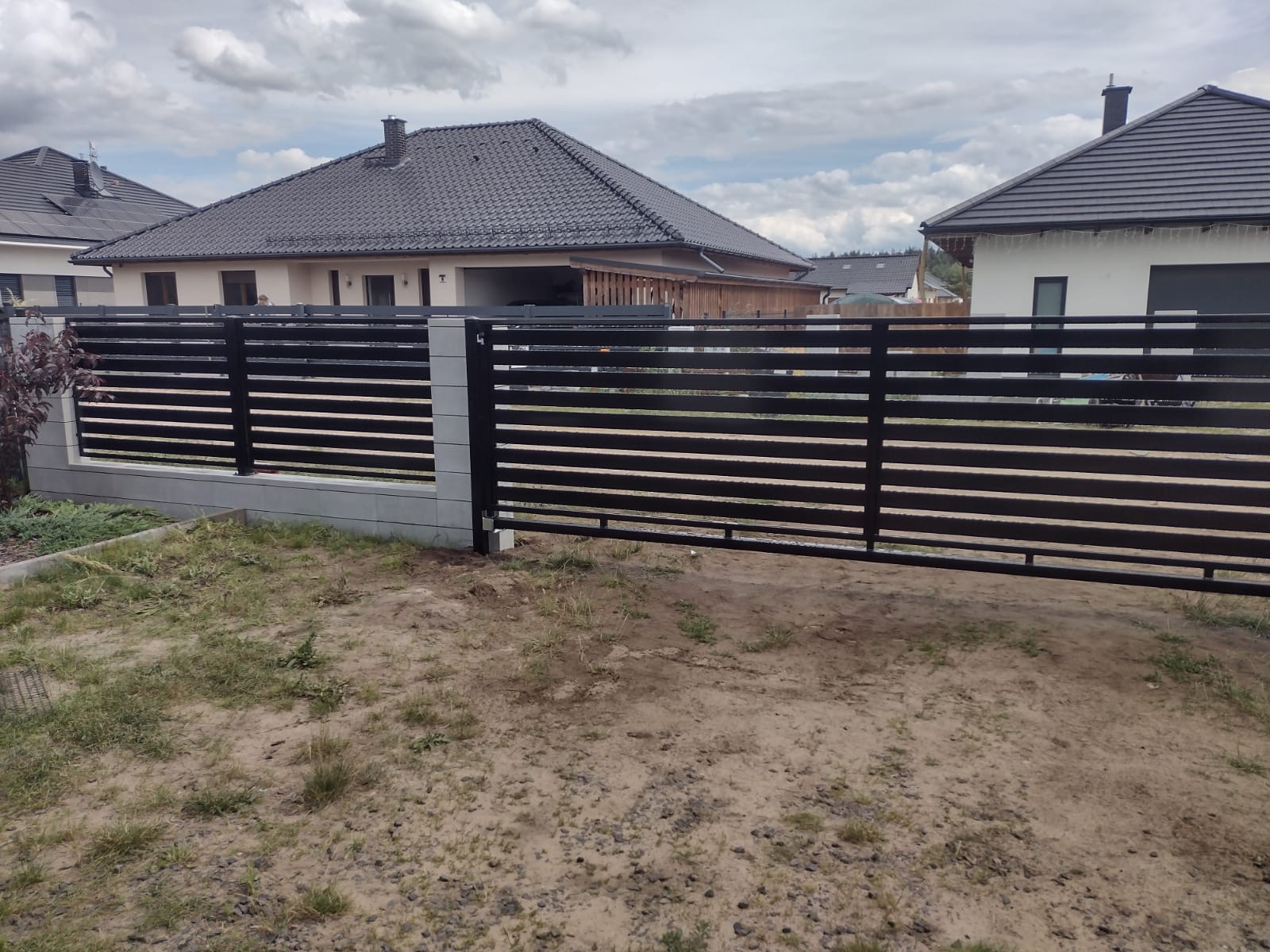 Nowoczesne ogrodzenie panelowe z poziomymi, czarnymi przęsłami i betonowymi słupkami na tle domów jednorodzinnych pod pochmurnym niebem.