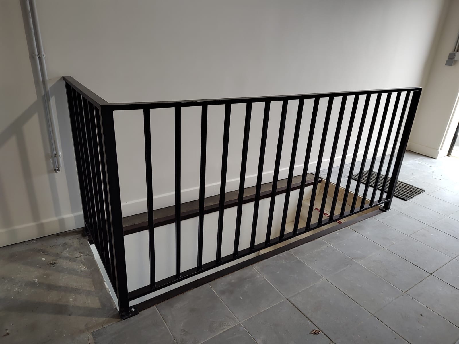 Czarne metalowe zabezpieczenie schodów piwnicznych z pionowymi szczebelkami, zamontowane na szarych płytkach podłogowych wewnątrz budynku.