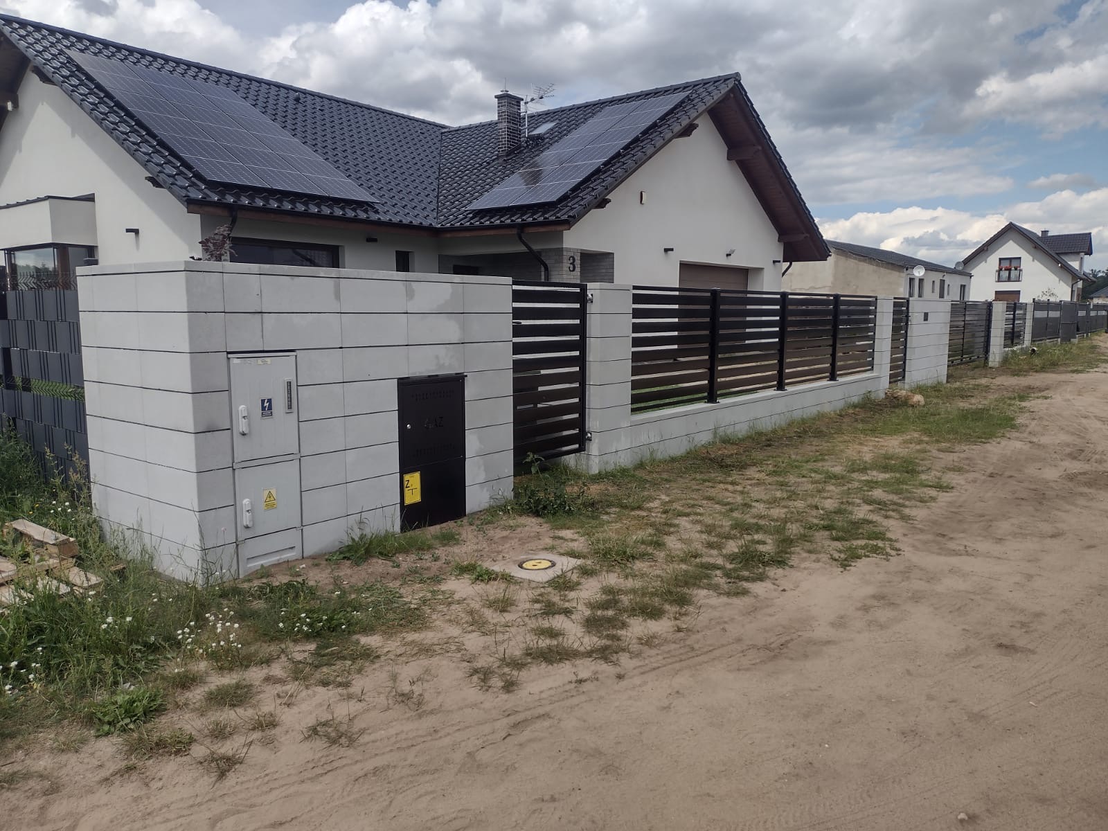 Nowoczesne ogrodzenie z betonowych bloczków i poziomych przęseł z ciemnego drewna, z szafkami elektrycznymi wkomponowanymi w konstrukcję, na tle domu z panelami słonecznymi.