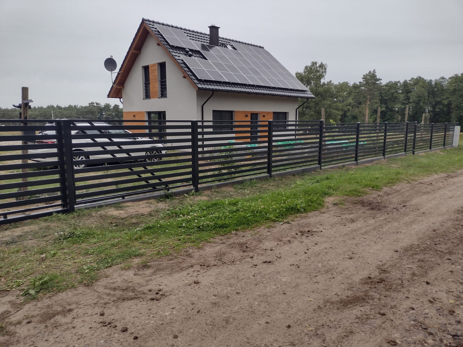 Nowoczesne, poziome przęsła z czarnych profili tworzące ogrodzenie posesji z domem jednorodzinnym z panelami słonecznymi na dachu.